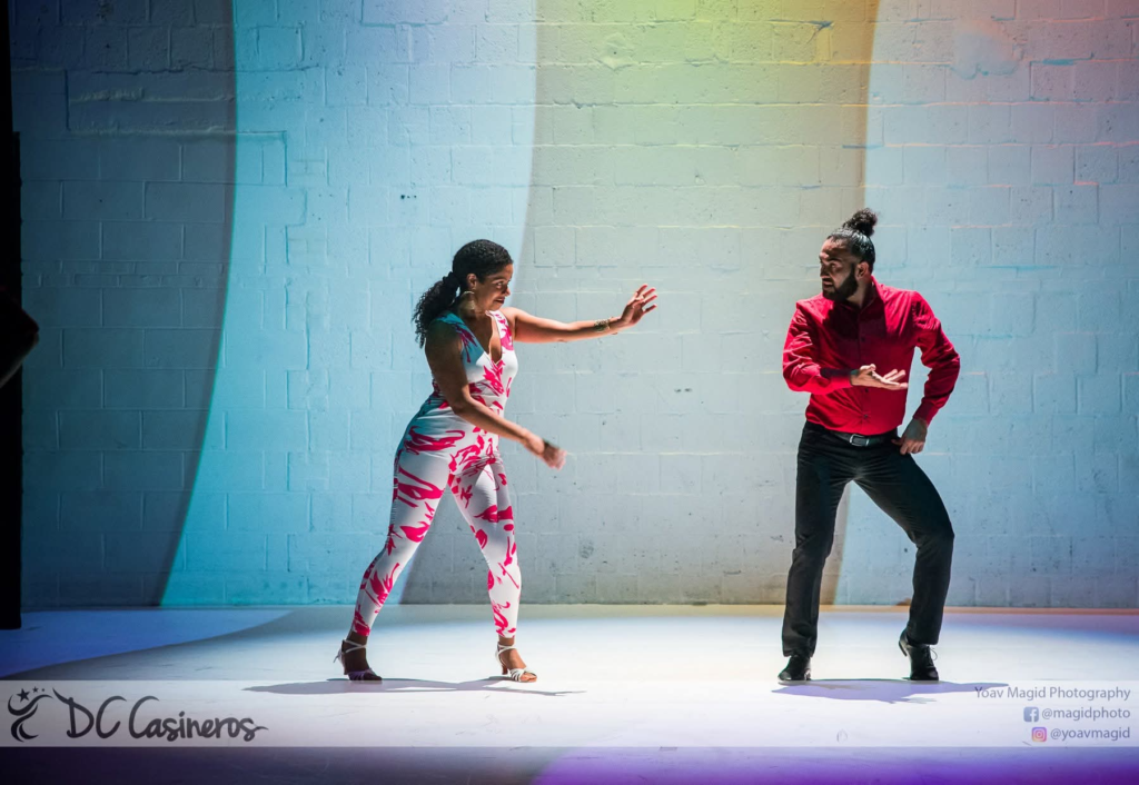 Two DC Casineros dancers performing together, wearing a floral jumpsuit and a red shirt