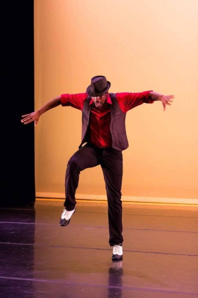 A DC Casineros performer in a hat, red shirt and dark vest dancing on stage.