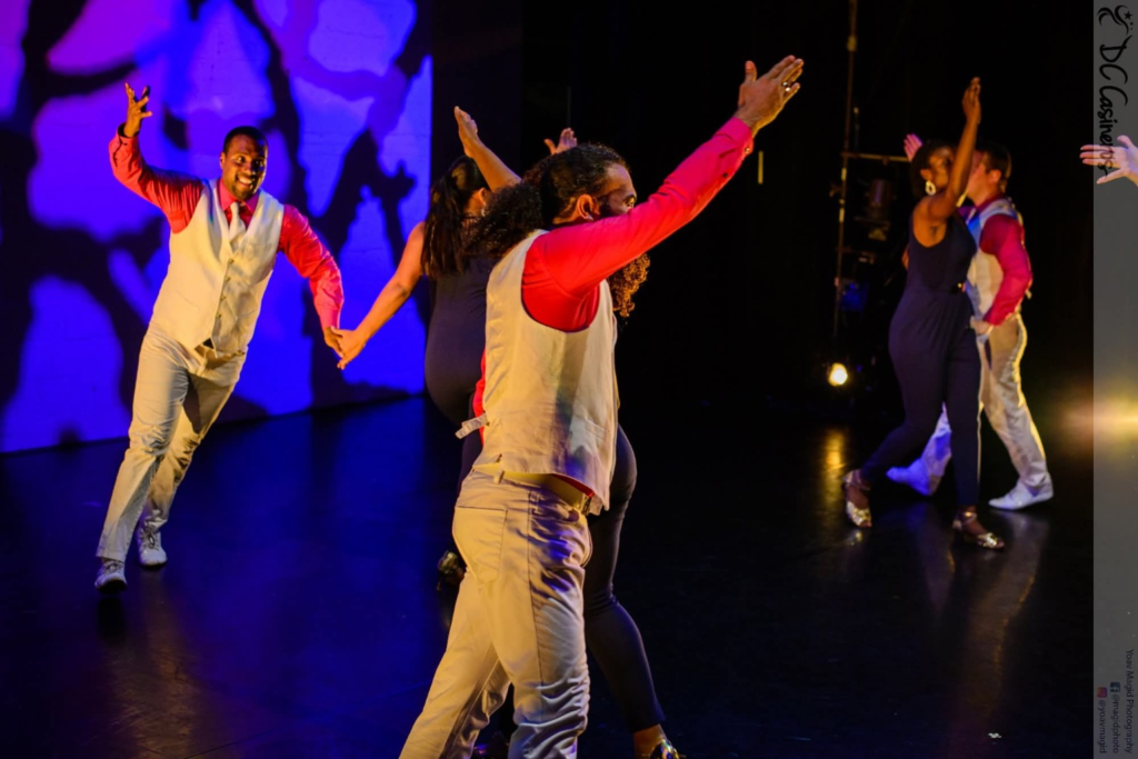 DC Casineros dancers performing in red shirts and pale vests under blue stage lights.